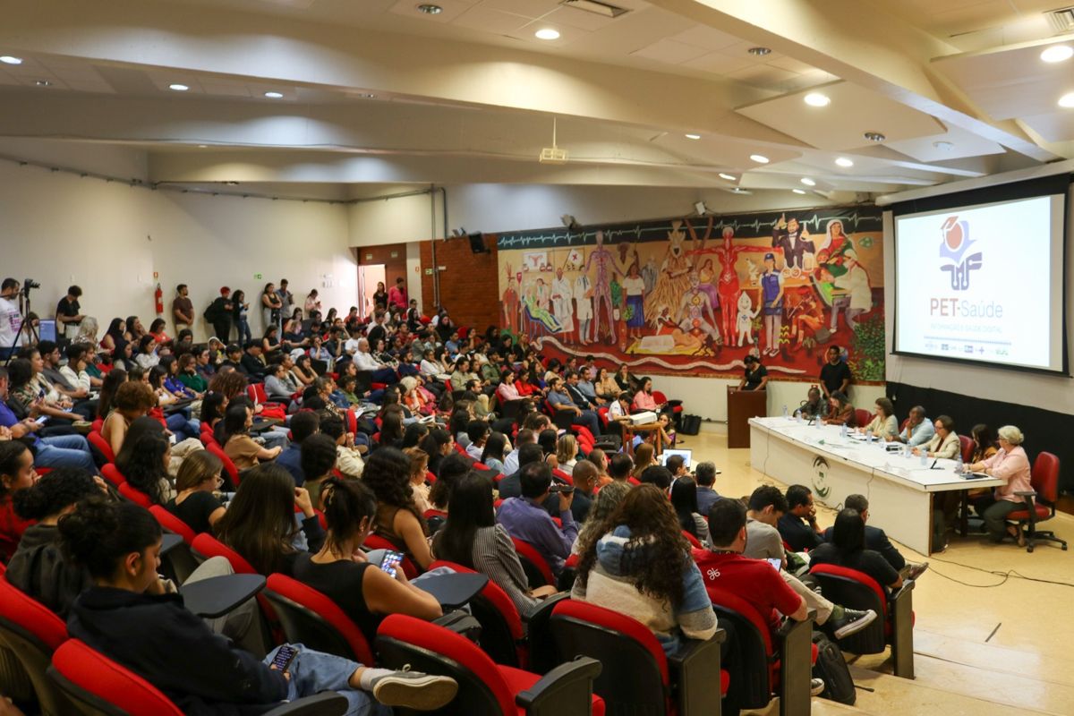 Auditório lotado em encontro do PET-Saúde Informação e Saúde Digital, com mesa de abertura e projeção do logo do programa.