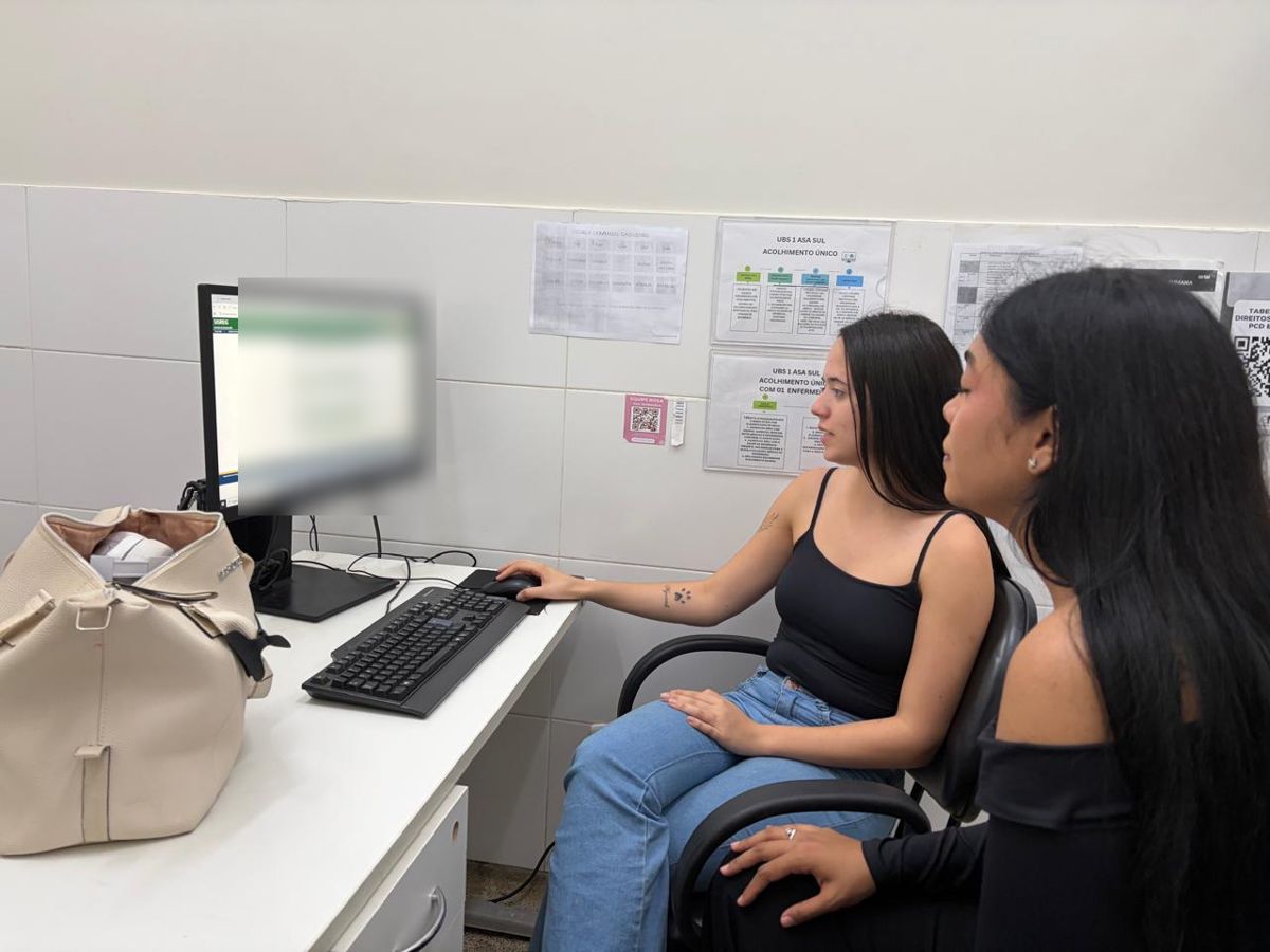 Duas integrantes do GT 12 em frente a um computador em sala da UBS 1 da Asa Sul, com cartazes de fluxo de acolhimento na parede.